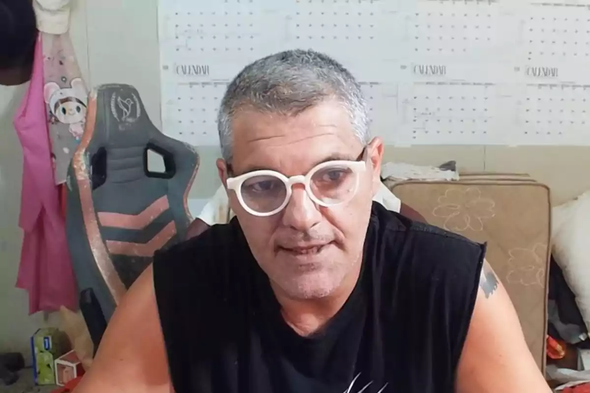 Frank Blanco con gafas blancas y camiseta negra está sentado frente a un calendario en la pared, con una silla de juego a su lado. Frank Blanco con gafas blancas y camiseta negra está sentado frente a un calendario en la pared, con una silla de juego a su lado.