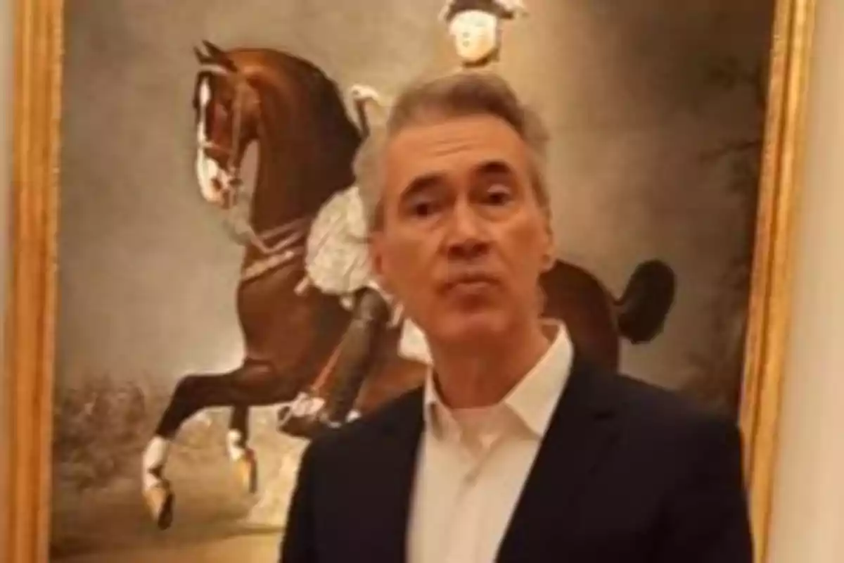 Hombre de cabello canoso con saco oscuro y camisa blanca posando frente a un cuadro de un jinete montando un caballo Hombre de cabello canoso con saco oscuro y camisa blanca posando frente a un cuadro de un jinete montando un caballo