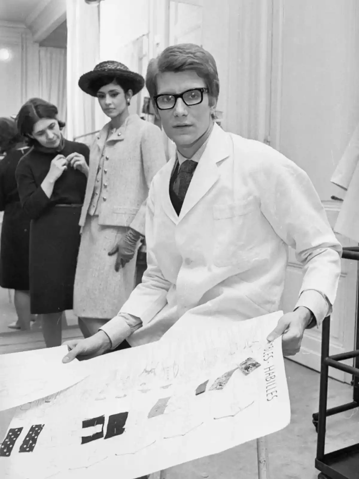 Un joven diseñador de moda con gafas y bata blanca sostiene un boceto mientras dos mujeres elegantemente vestidas observan en el fondo Un joven diseñador de moda con gafas y bata blanca sostiene un boceto mientras dos mujeres elegantemente vestidas observan en el fondo