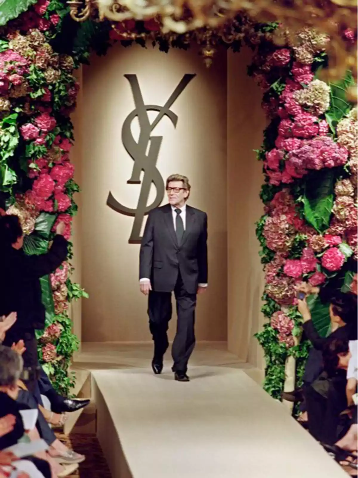 Hombre con traje oscuro caminando por una pasarela decorada con flores y el logo de YSL al fondo mientras el público aplaude Hombre con traje oscuro caminando por una pasarela decorada con flores y el logo de YSL al fondo mientras el público aplaude