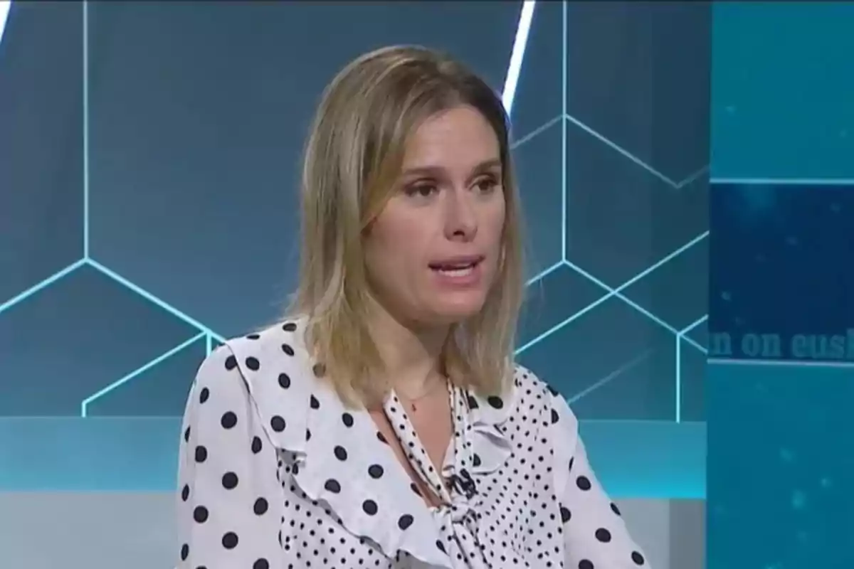Una mujer con blusa de lunares habla en un estudio de televisión con fondo geométrico. Una mujer con blusa de lunares habla en un estudio de televisión con fondo geométrico.