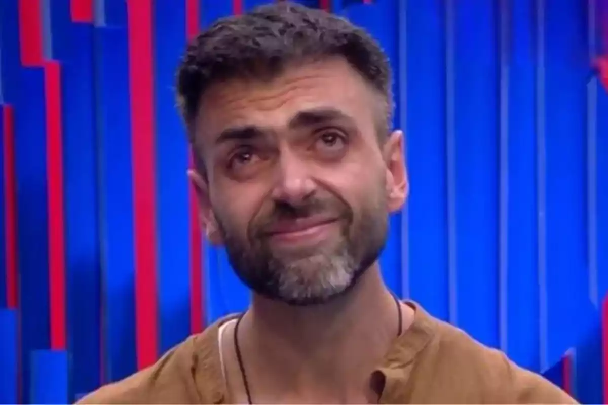 Hombre con barba y cabello corto mirando hacia arriba con expresión emotiva y fondo azul con líneas rojas. Hombre con barba y cabello corto mirando hacia arriba con expresión emotiva y fondo azul con líneas rojas.